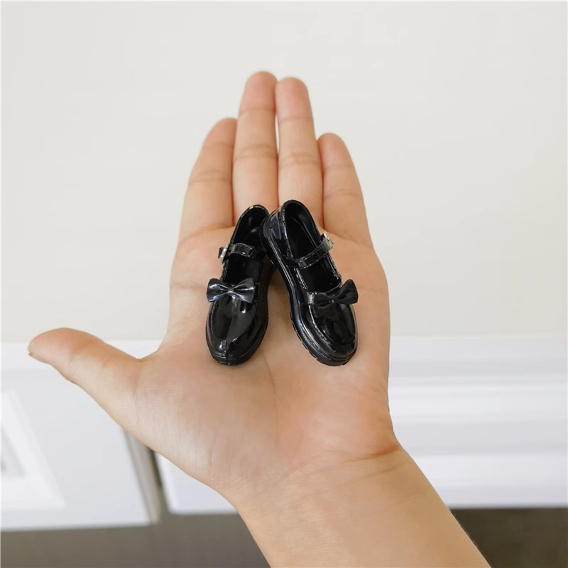 Actual black 1:6 figure bow-strap miniature shoe pair displayed in a hand for scale; hand not included